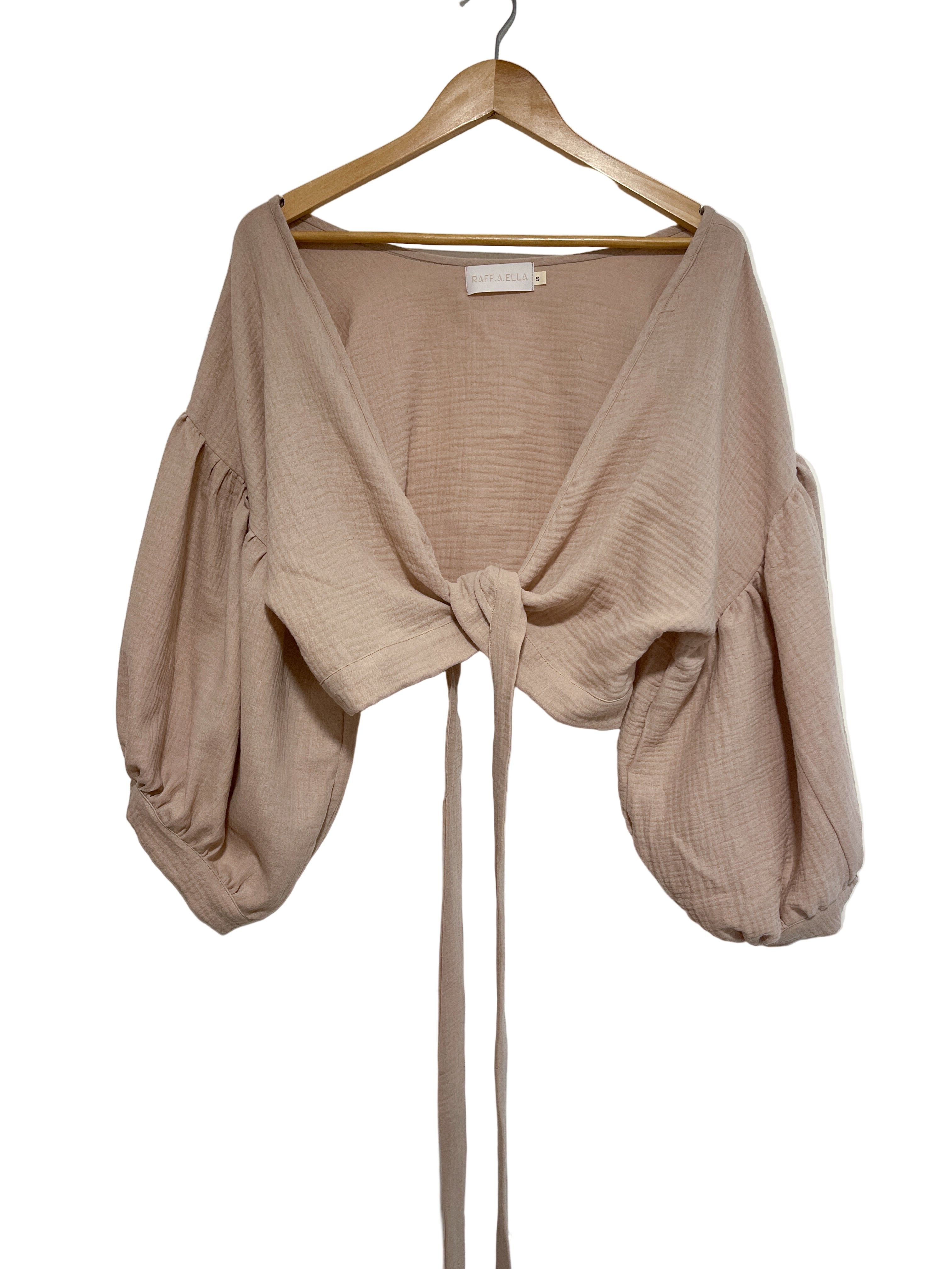 A beige 'Persephone' wrap top with balloon sleeves and a self-tie waistband, displayed on a wooden hanger against a white wall.